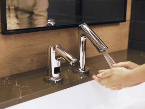 Persona lavándose las manos bajo un grifo con sensor sin contacto de acero inoxidable, junto a un dispensador de jabón automático sobre un lavabo moderno.
