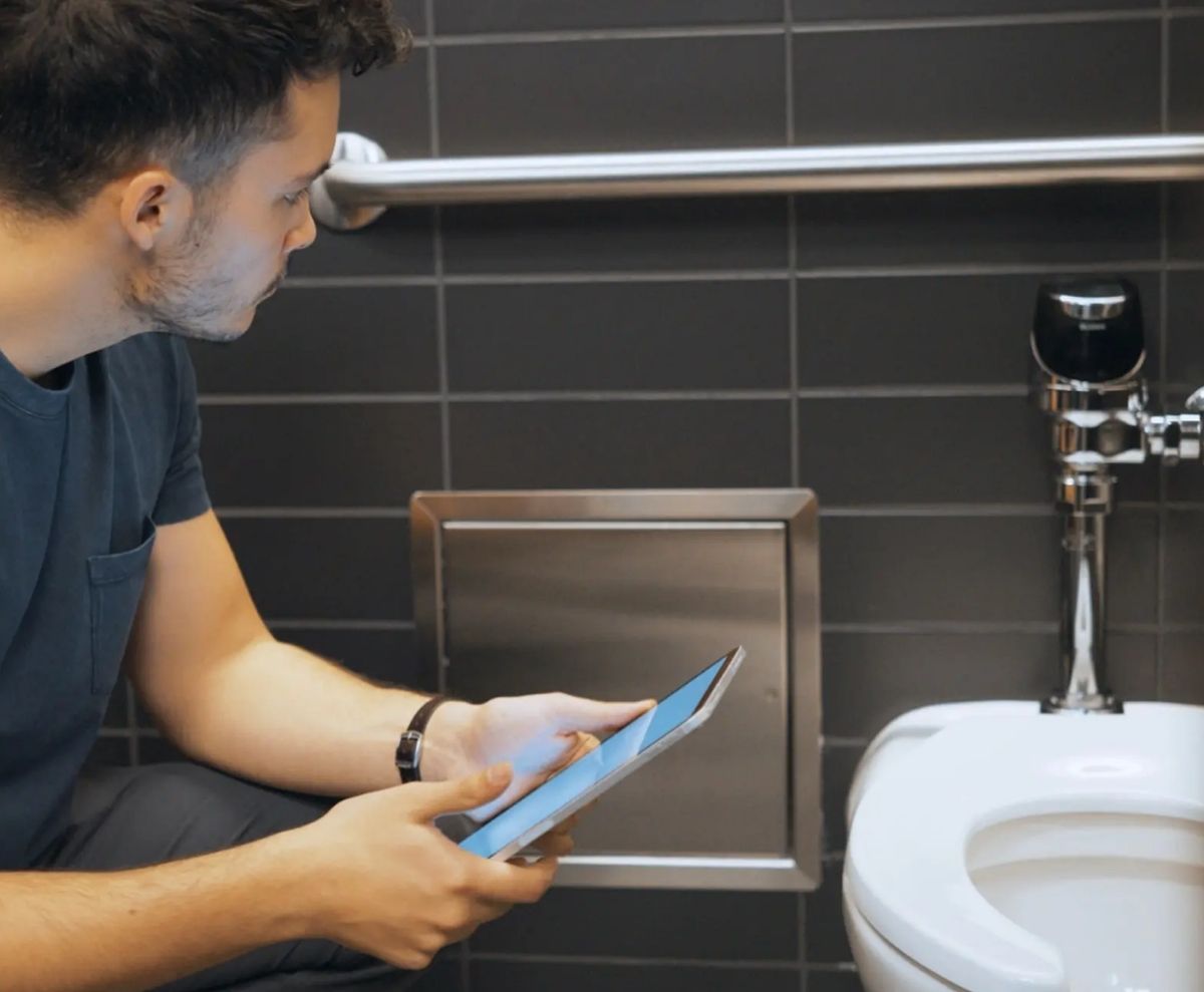 Hombre usando una tableta mientras inspecciona un sanitario con barra de apoyo en un baño de azulejos oscuros.
