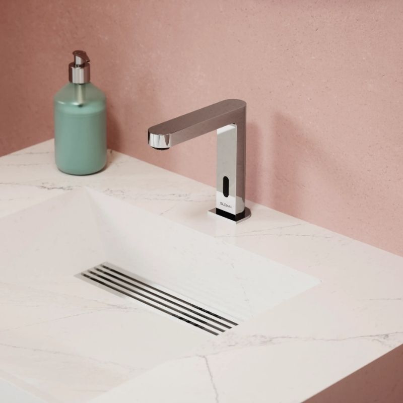 Sloan automatic chrome faucet with clean, angular design, mounted on a white marble countertop with a pink wall and green soap dispenser.