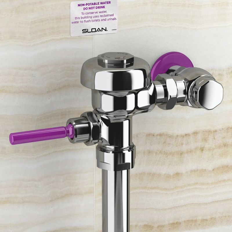 Sloan manual flushometer in polished chrome finish with purple handle and rear cover, indicating use of reclaimed water, installed against a beige marble wall.