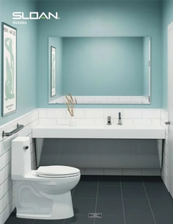 hotel restroom with a modern and welcoming design. Soft blue walls combine with white tiles and a wide sink featuring Sloan faucets. The space conveys cleanliness, comfort, and functionality—perfect for a hospitality setting.
