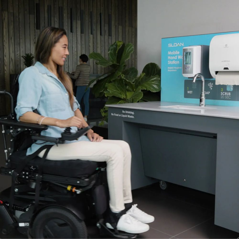Mujer en silla de ruedas usando una estación móvil de lavado de manos accesible, equipada con un dispensador automático y un módulo de toallas de papel, en un entorno moderno e inclusivo.