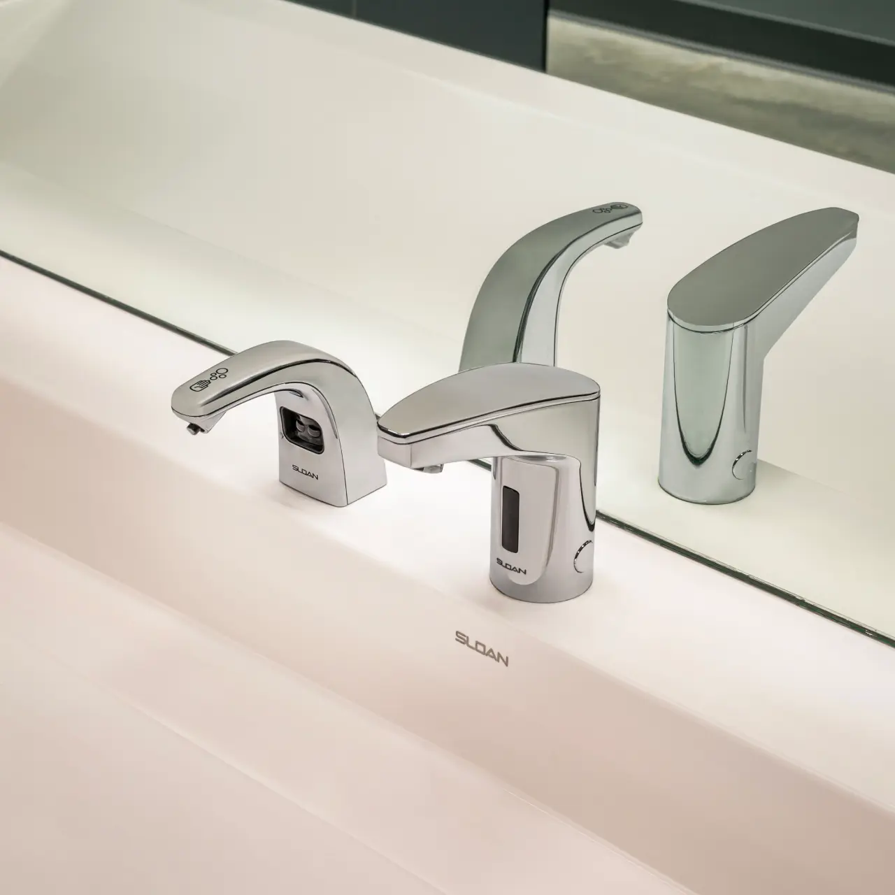modern sink featuring two Sloan automatic chrome faucets integrated into a clean white surface. The mirror reflection enhances symmetry and the sense of hygiene, while the white lighting highlights the metallic shine and the technological elegance of the design.