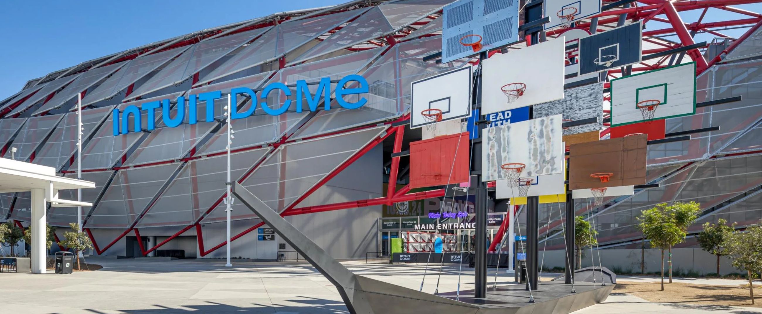 Exterior view of the Intuit Dome featuring its distinctive red and gray steel structure, with an artistic basketball backboard installation in front of the main entrance.