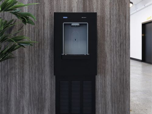 Dispensador de agua empotrado negro en un pasillo moderno con paredes de textura de madera y una planta decorativa cercana.