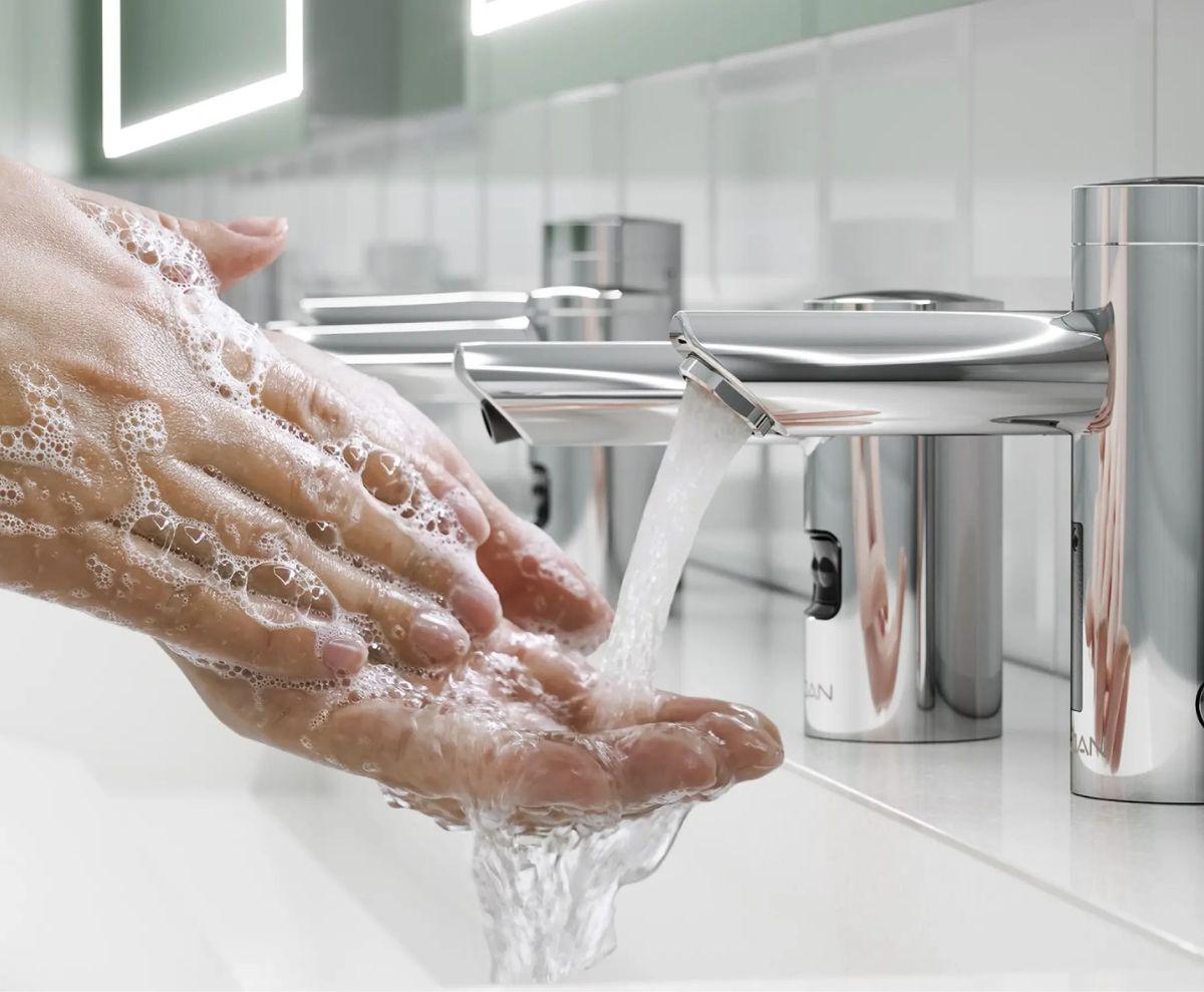 Persona lavándose las manos con jabón bajo un grifo automático de acero inoxidable en un lavabo moderno que promueve la higiene y el bienestar.