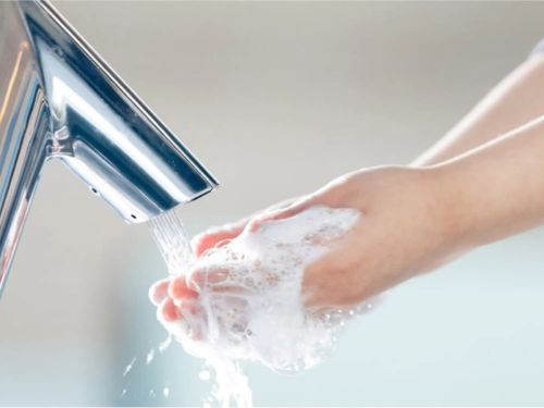Persona lavándose las manos con jabón bajo un grifo automático de acero inoxidable, representando prácticas de higiene.