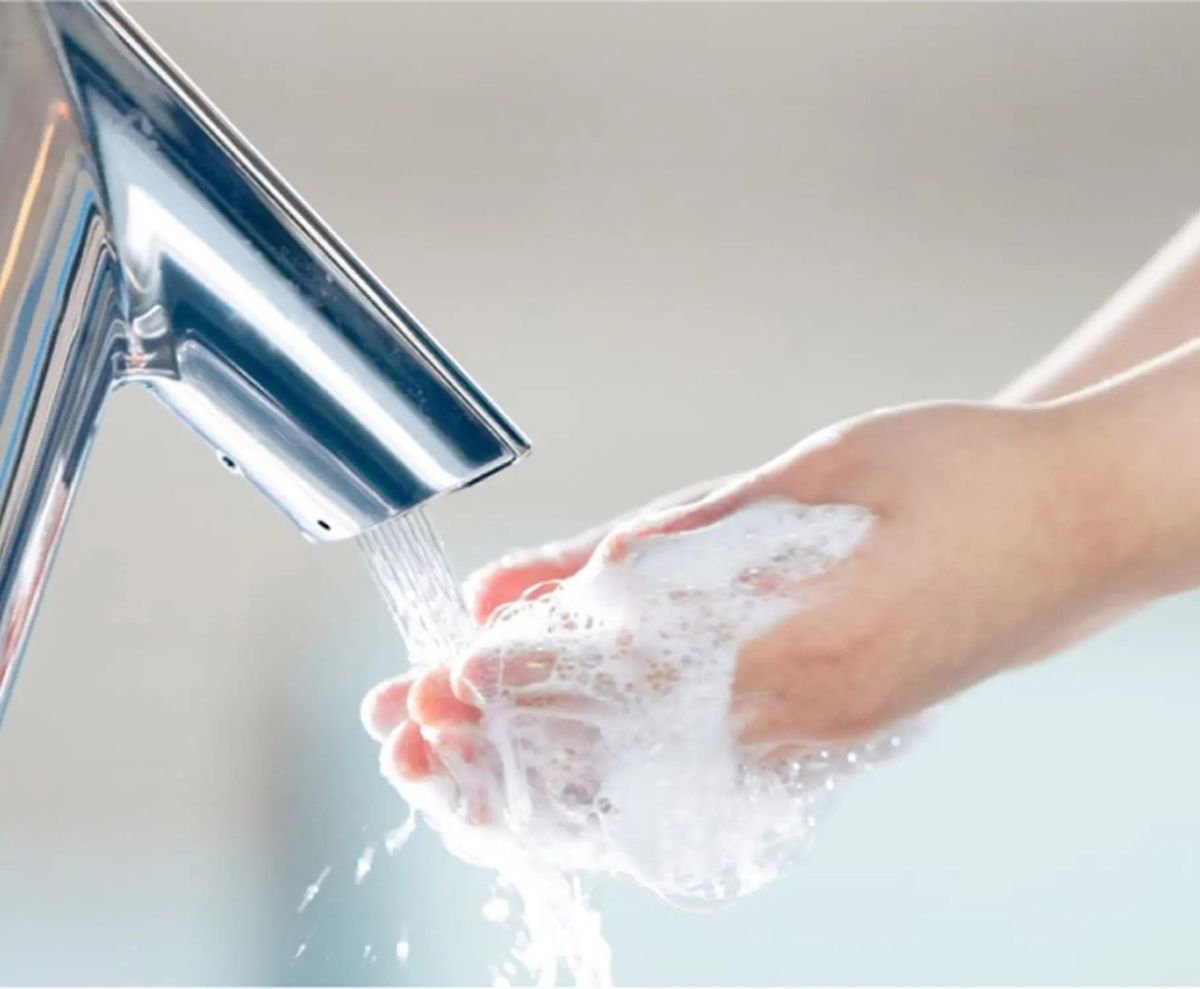 Persona lavándose las manos con jabón bajo un grifo automático de acero inoxidable, representando prácticas efectivas de lavado de manos e higiene.
