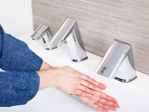 Persona secándose las manos bajo un secador automático integrado, junto a un grifo sin contacto y un dispensador de jabón sobre un lavabo moderno.