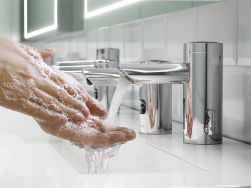 Persona lavándose las manos con jabón bajo un grifo automático de acero inoxidable en un baño moderno, promoviendo la higiene y la seguridad.