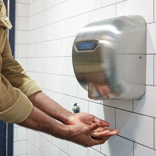 Person drying their hands with a stainless steel Sloan XLERATOR hand dryer mounted on a white tiled wall.