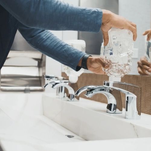 Person refilling a Sloan soap dispenser installed next to an automatic faucet in a commercial sink.