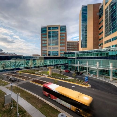 Exterior view of a modern hospital building with glass walkways and a bus passing by the entrance.