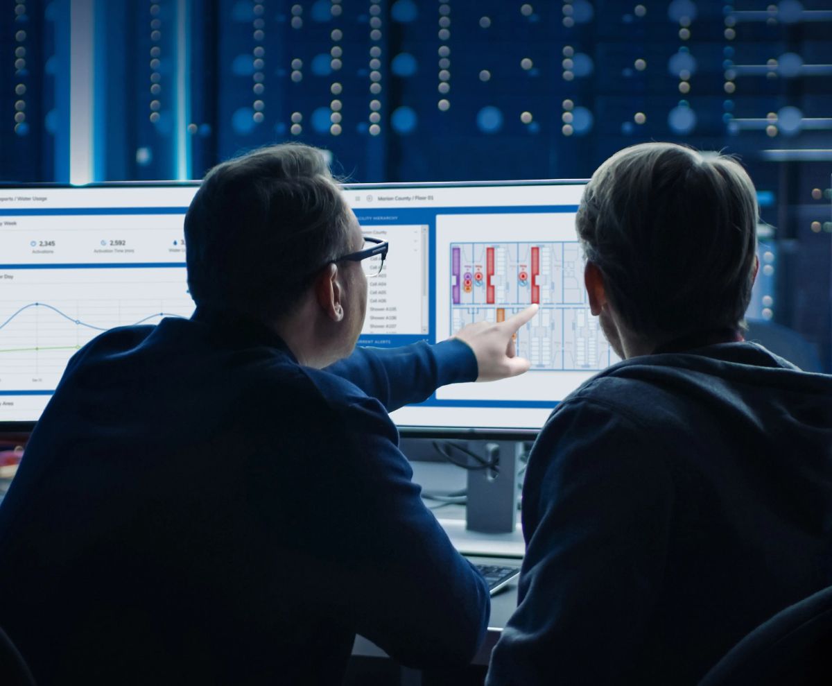 Dos personas analizando datos y diagramas en monitores de computadora, revisando información técnica o de desempeño en un entorno de control digital.