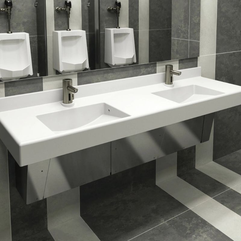 Lavabo doble de superficie sólida blanca con grifos automáticos Sloan, instalado frente a una pared con azulejos grises y mingitorios al fondo.