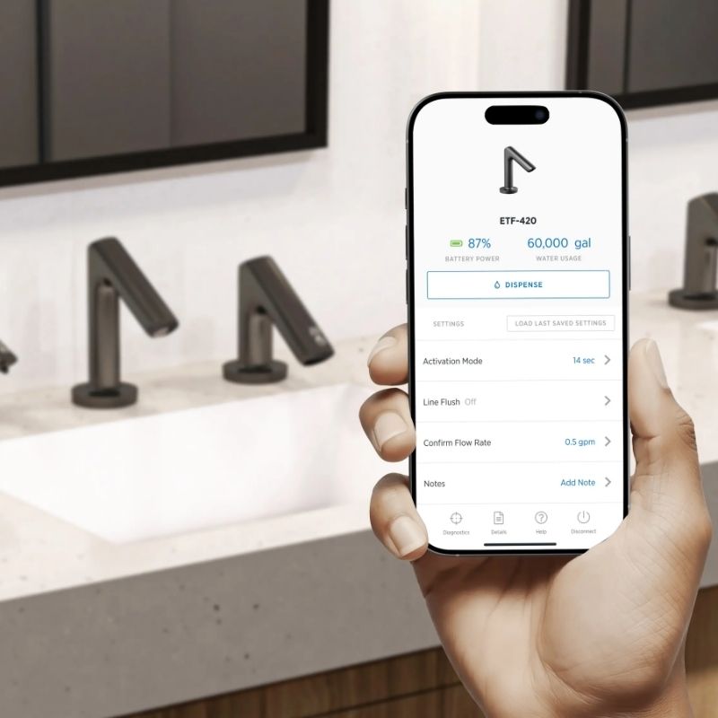 Hand holding a smartphone displaying the Sloan Connect app with data and settings for a smart faucet, in front of sinks with Sloan sensor faucets in matte black finish.