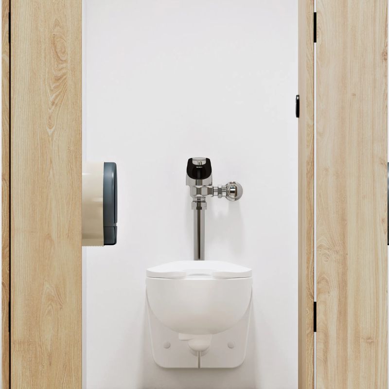 Wall-mounted toilet with Sloan automatic flushometer in a modern restroom stall featuring light wood doors and a white wall.