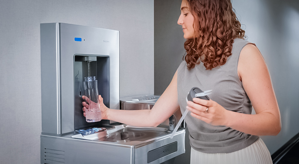 Woman refilling a reusable bottle at a Sloan stainless steel water dispenser in a modern hallway.