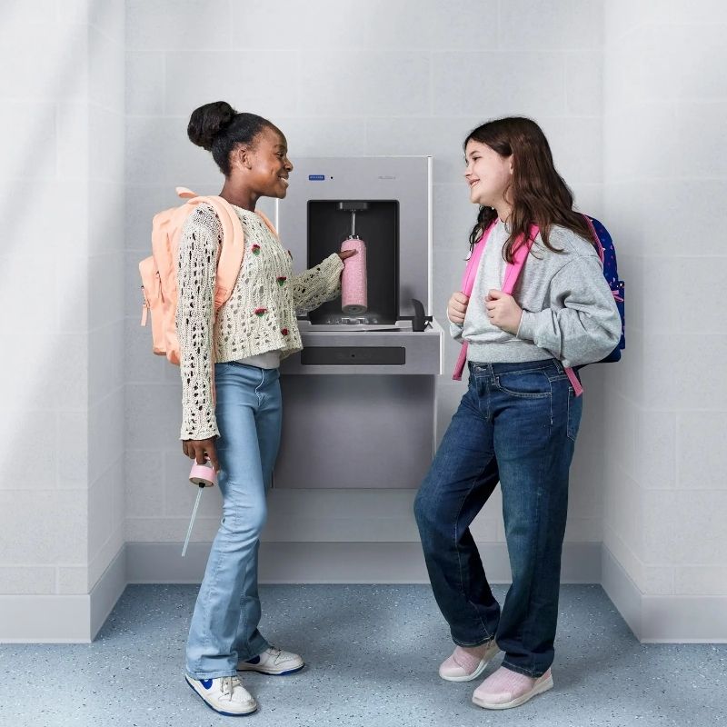 Two students filling a pink bottle at a Sloan water bottle filler in a school hallway with blue lockers.