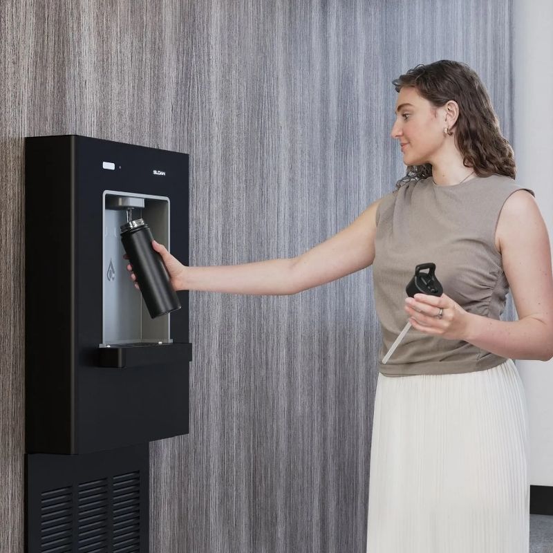 Woman refilling a black reusable bottle at a Sloan wall-mounted water dispenser with a wood-textured background.