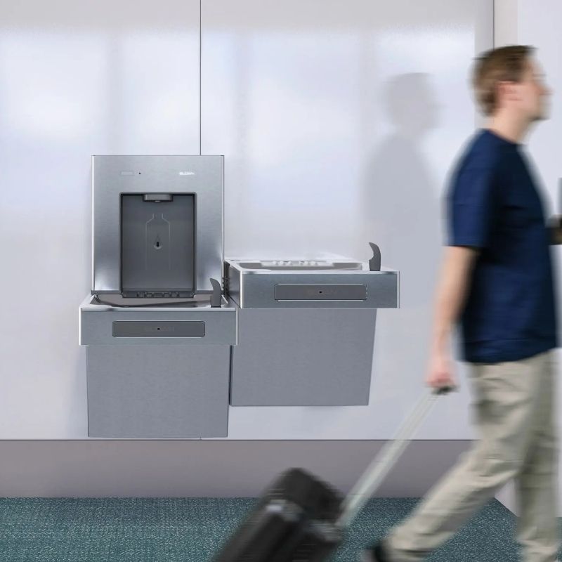 Stainless steel Sloan wall-mounted water dispenser and drinking fountain in a public space, with a person walking by pulling luggage.