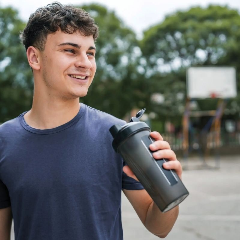 Smiling young man holding a reusable water bottle outdoors near a basketball court.