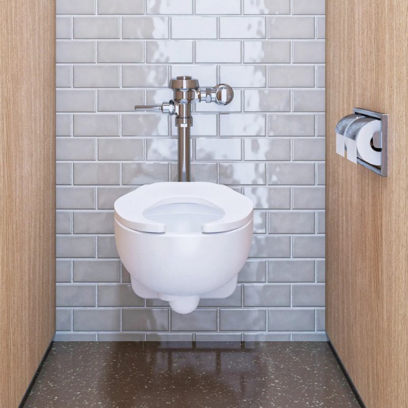 Wall-mounted toilet with Sloan manual flushometer in polished chrome finish, installed in a stall with gray tiled walls and light wood partitions.