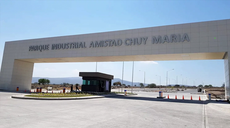 the main entrance of the Amistad Chuy María Industrial Park, a modern structure with a large light-colored gateway. At the center stands a security booth surrounded by wide, well-paved roads, with a semi-arid landscape visible in the background.