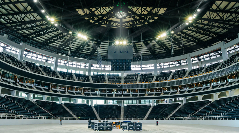 the interior of a modern sports arena with spacious seating and a steel-structured roof. A central screen bearing the Sloan logo hangs above, while the overhead lighting emphasizes the scale and architectural design of the venue.