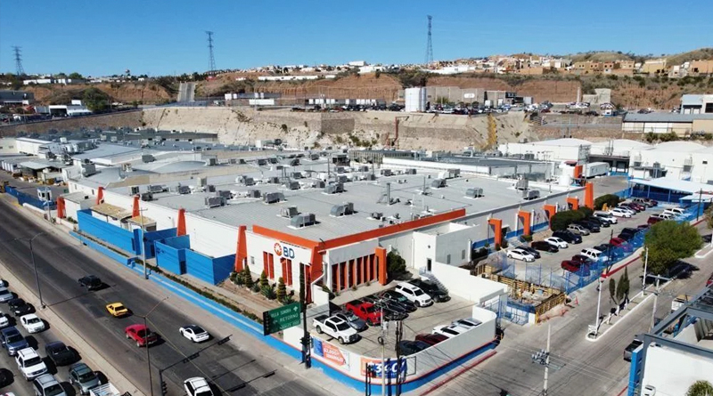 an aerial view of a modern industrial complex featuring large metal-roofed warehouses and an exterior design in white, blue, and orange tones. In the foreground, there are well-organized parking areas and a main avenue with light traffic, while in the background, urbanized hills and supporting structures complete the setting.