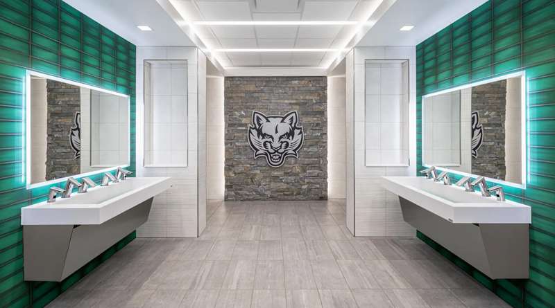 contemporary high-design restroom featuring two opposing double sinks, each with chrome automatic faucets and backlit mirrors. The side walls are clad in glossy green tiles that contrast with the light gray floor and central stone walls. At the back, a stone accent wall with a feline logo adds an institutional and athletic touch. Linear ceiling lighting enhances the symmetry, creating a modern, clean, and visually striking environment.