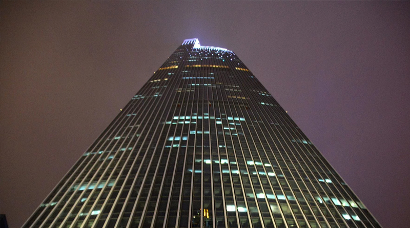 an impressive skyscraper captured from its base, highlighting its modern design with vertical lines that emphasize its height. The illuminated windows contrast against the night sky, creating an urban and elegant atmosphere.
