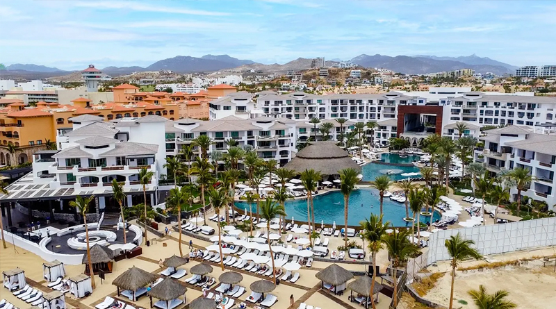 panoramic view of the Cabo Azul Resort Hotel, a luxury beachfront complex located in San José del Cabo, Mexico. The resort features contemporary architecture with tropical accents, expansive pools surrounded by palm trees and sun loungers, and a distinctive central palapa roof that serves as the visual centerpiece of the property.