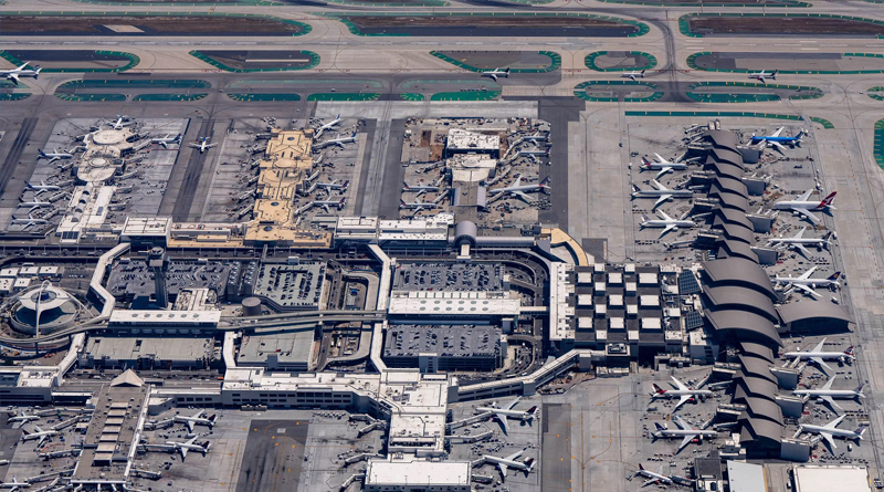 an aerial view of a large international airport, with multiple terminals connected by walkways and aprons filled with airplanes from various airlines. Runways and taxiways extend in the background, while the aircraft parking areas and access roads are clearly organized. The aerial perspective highlights the scale and complexity of the airport’s infrastructure.