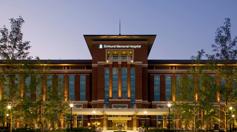 the main facade of Elmhurst Memorial Hospital, a modern brick building with tall vertical windows and warm lighting that highlights its architecture at dusk. The landscaped surroundings and symmetrical walkways create an atmosphere of calmness and professionalism.
