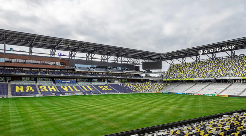 the interior of GEODIS Park in Nashville, home of Nashville SC. The stadium features a modern design with large covered stands, seats in shades of blue, yellow, and gray, and a meticulously maintained grass field. The architecture combines exposed steel and clean lines, reflecting a contemporary industrial style.