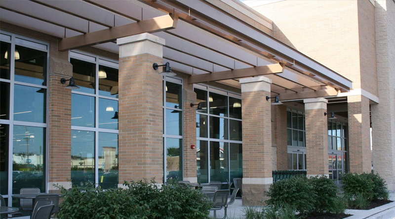 the facade of a modern commercial building featuring a brick structure, large windows, and columns supporting a metal canopy. The design incorporates landscaped areas and outdoor seating, creating a pleasant and functional space for customers and visitors.