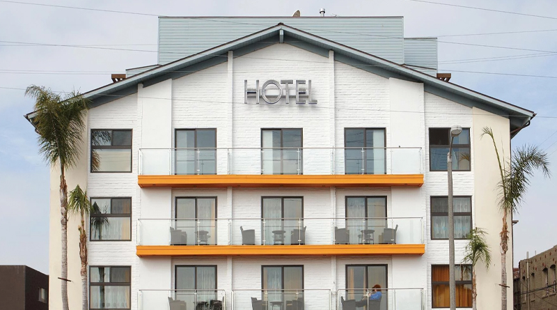 Hotel Erwin, an iconic property located in Venice Beach, Los Angeles. The building features a modern, laid-back design with white walls, glass balconies, and bright orange accents that capture the lively beach atmosphere. Palm trees frame the exterior, enhancing the coastal California vibe, while the bold metallic “HOTEL” sign underscores its minimalist and contemporary style.