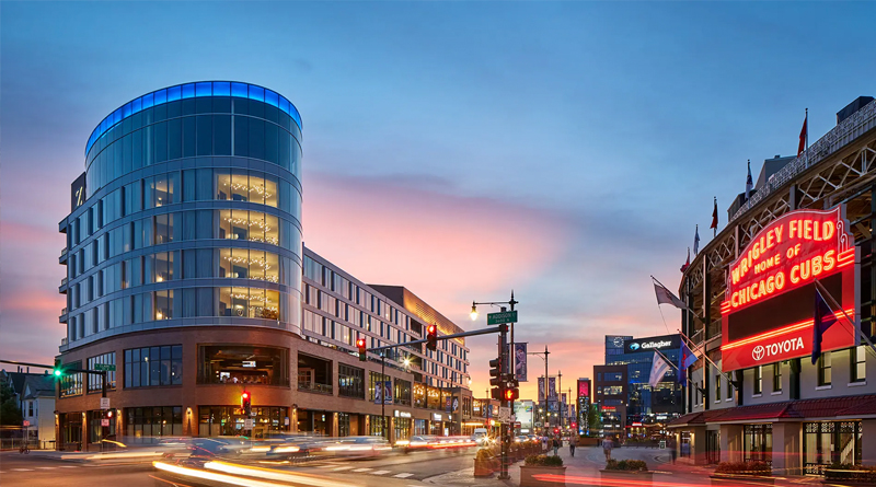 Hotel Zachary, located across from the iconic Wrigley Field, home of the Chicago Cubs. The building blends contemporary architecture with classic brick elements, complementing the stadium’s historic character. Its curved glass façade glows at sunset, reflecting the lively urban atmosphere of Chicago’s Lakeview neighborhood.