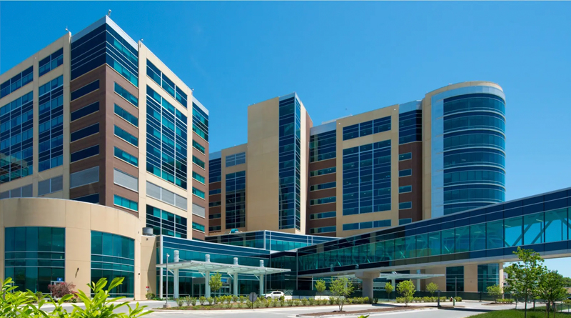 a modern hospital complex with large glass façades and contemporary architectural design. The building features beige and brown tones with blue-tinted windows that reflect the sky, conveying a sense of cleanliness, innovation, and efficiency. In the foreground, landscaped greenery and a driveway lead to the main entrance with a glass canopy.