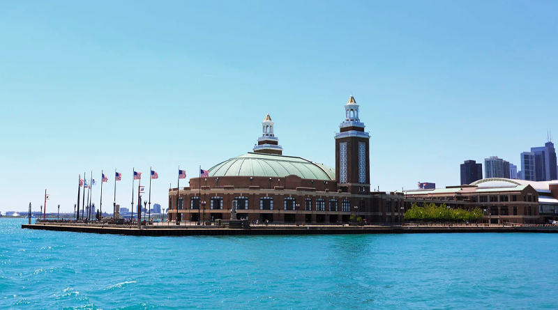 Navy Pier in Chicago, Illinois — one of the city’s most iconic tourist landmarks. The main building, with its green dome and twin brick towers, extends over Lake Michigan, surrounded by a pier lined with multiple U.S. flags waving in the breeze. The turquoise water and clear sky highlight the architectural elegance of the venue, which blends history and modernity as it hosts events, restaurants, and cultural attractions.