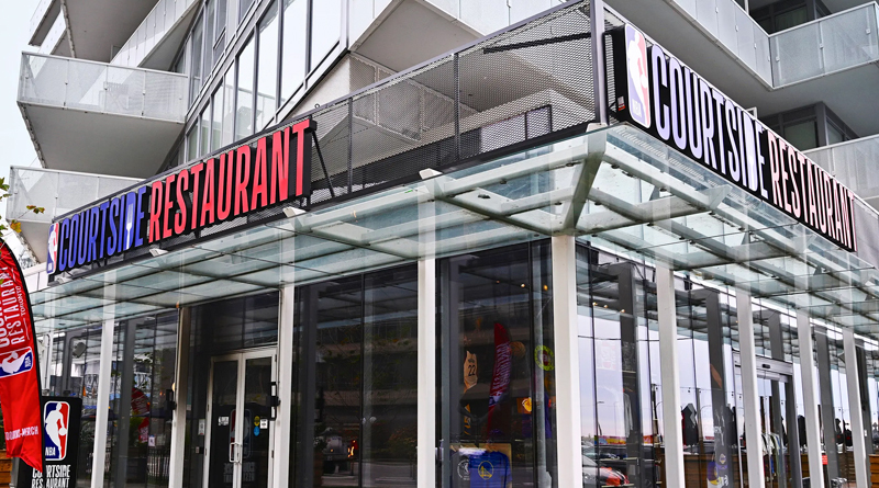 the exterior of the NBA Courtside Restaurant, a modern, NBA-themed dining venue. The building features glass and metal architecture with clean lines, highlighted by a bold sign displaying the league’s official logo and the restaurant name. The overall design conveys an urban, contemporary feel, perfect for basketball fans seeking a sports-inspired dining experience.