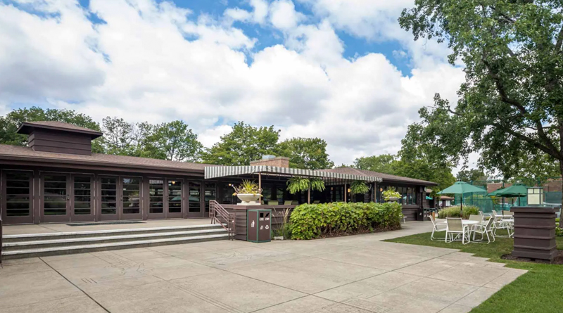 River Forest Tennis Club, a classic American-style building with a low, horizontal structure, large windows, and dark wood finishes. The surroundings feature well-maintained greenery, white outdoor furniture, and a paved area leading toward the tennis courts.