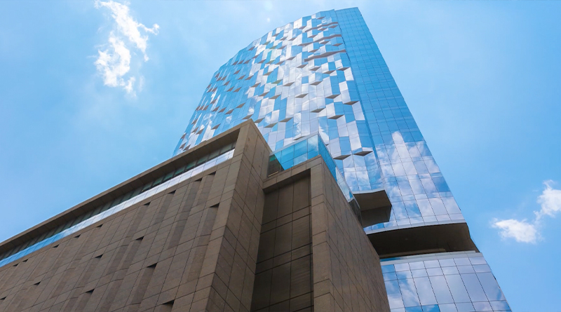 modern skyscraper with a reflective glass façade that mirrors the blue sky and clouds. At the base, brown-toned structures contrast with the metallic shine of the main tower, highlighting its contemporary and elegant design.