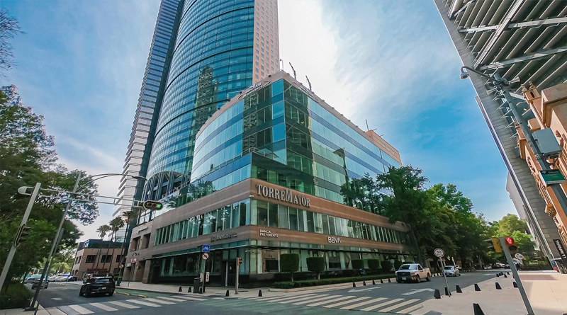 Torre Mayor, a modern skyscraper located in Mexico City. Its façade combines reflective glass and metal structures, standing out for its sleek design and impressive height. At the base, office spaces and bank branches are visible, while the surrounding urban environment conveys an orderly, contemporary atmosphere under a clear sky.
