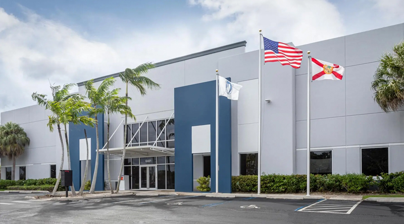 the main façade of William R. Nash’s corporate offices, a Florida-based company specializing in mechanical, plumbing, and HVAC engineering solutions. The building features modern industrial architecture with light gray and blue panels, large windows, and a glass-fronted central entrance that brings in natural light. In front, three flags fly —those of the United States, the State of Florida, and the company flag— symbolizing its professional identity and local presence.