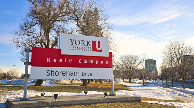 the main entrance of York University’s Keele Campus in Toronto, Canada. The red-and-white sign, featuring the university’s logo, indicates the Shoreham Drive entrance — one of the key access points to the campus. The snow-covered ground and bare trees evoke the region’s characteristic winter season.